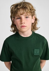 Grøn bomulds T-shirt med korte ærmer, der har et firkantet gummilogo-patch på venstre bryst. Glat tekstur med en rund halsudskæring.