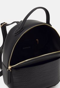 Sac à dos noir de forme arrondie, en tissu lisse avec une base texturée. Présente une fermeture éclair dorée et une poche intérieure avec un logo.