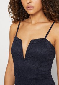 Vestido de renda azul marinho com decote em V profundo e alças finas. O tecido apresenta um padrão floral e uma superfície texturizada.