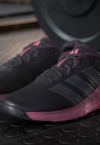 Μαύρα και σκούρα ροζ αθλητικά παπούτσια Adidas με τρεις μαύρες ρίγες, τοποθετημένα πάνω σε δάπεδο γυμναστηρίου κοντά σε πλάκα βαρών.