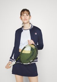 Navy bomber jacket with striped collar, white cropped top with vertical stripes, and navy skirt. Holding a tennis racket with a green grid pattern.