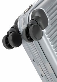 Valigia rigida argentata con design a righe verticali, dotata di doppie ruote nere e chiusura con zip lungo il lato.