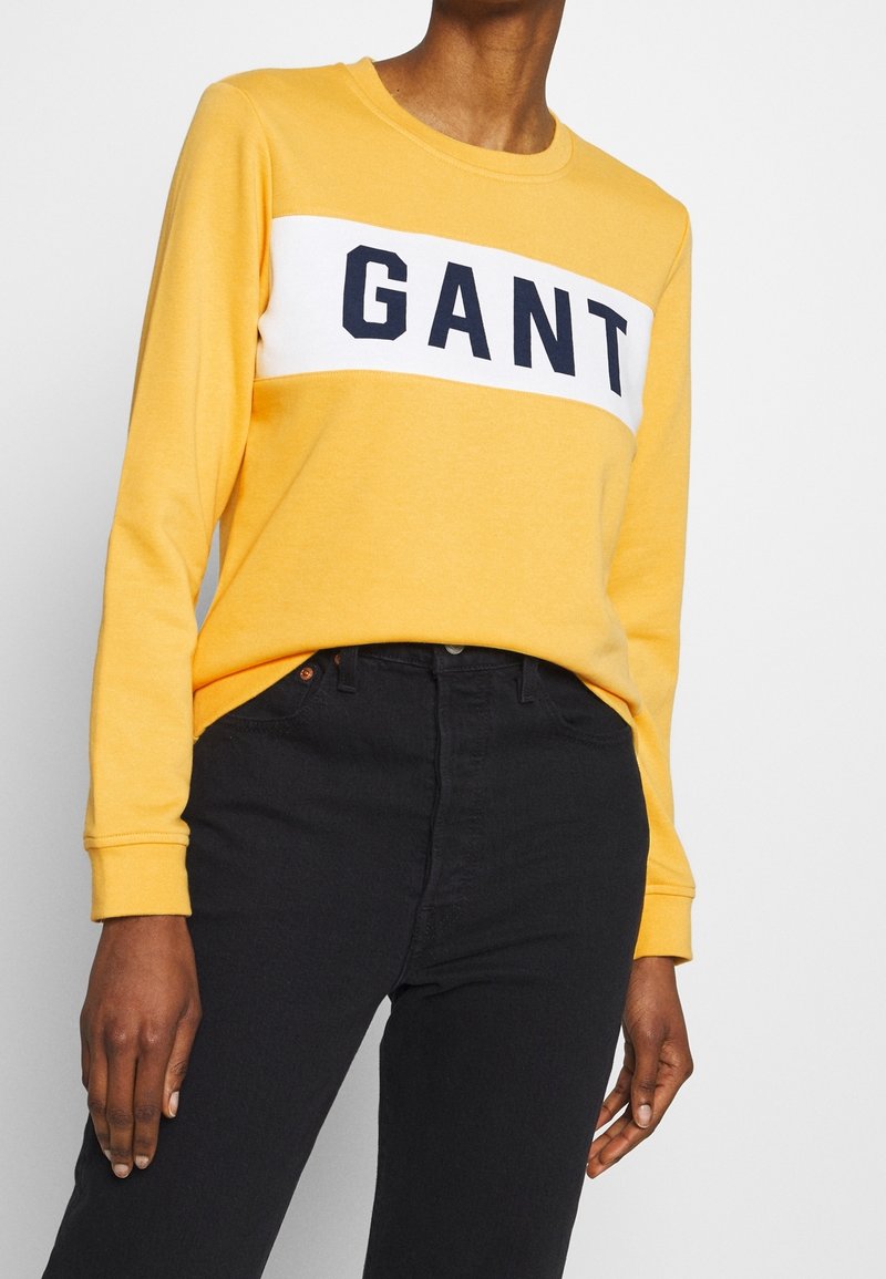 Geel sweatshirt met een witte horizontale panel waarop het woord "GANT" in marineblauw staat. Lange mouwen, ronde hals, gecombineerd met zwarte jeans.