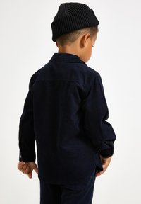 Granatowa koszula z korduroyu z długimi rękawami, kołnierzykiem i detalami mankietów na guziki. Noszona z czarną, prążkowaną czapką beanie. Widok z tyłu.