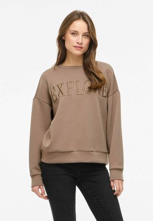 Junge Frau mit langen braunen Haaren, die einen braunen Pullover mit dem Schriftzug "EXPLORE" und schwarze Hosen trägt, steht vor einem weißen Hintergrund.