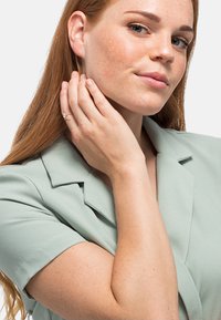 Hellgrüne Kurzarmbluse mit Kragen, kombiniert mit einem zarten goldenen Ring an ihrer Hand. Glatte Stoffoberfläche und natürliche Hauttöne.
