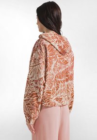 Frau mit langen dunklen Haaren, die einen lockeren Kapuzenpullover in Orange und Creme mit Muster sowie hellrosa Hose trägt, steht vor einem weißen Hintergrund.