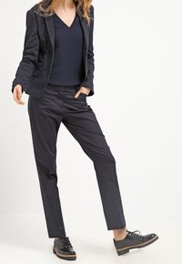 Kvinna i svart blazer med prickar, svart V-ringad topp, svarta skräddarsydda byxor och svarta blanka oxfordskor, stående på vit golv.