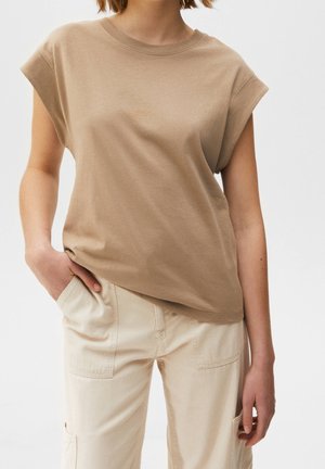 Femme portant un haut beige ample sans manches et un pantalon beige clair avec des poches, debout avec une main dans une poche devant un fond blanc.