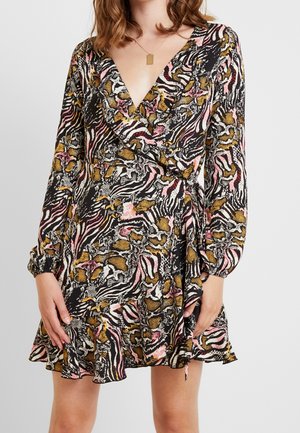 Femme portant une robe portefeuille à manches longues et col en V avec un ourlet volanté, présentant un imprimé animal abstrait multicolore en noir, blanc, rose et tons dorés.