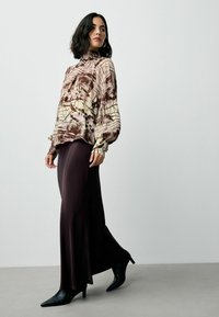 Langmouw blouse met patroon in crème en bruin, gecombineerd met een vloeiende donkerbruine rok en puntige zwarte hakken. Glad textuur aanwezig.