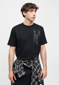 Νεαρός άνδρας κοιτάζει δεξιά φορώντας μαύρο T-shirt New Balance Athletics και μαύρο-λευκό καρό πουκάμισο δεμένο γύρω από τη μέση του, μπροστά σε λευκό φόντο.