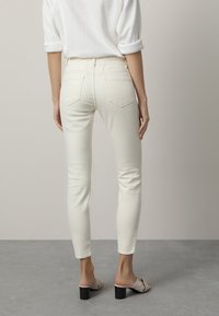 Femme portant un jean slim-fit blanc cassé, un top blanc à manches longues retroussées et des sandales à talons beiges, se tenant sur un sol gris.
