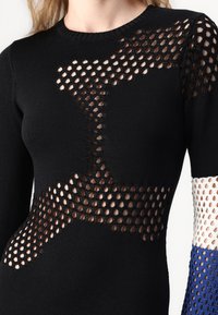 Svart stickad tröja med nätsektioner och diagonala skärningar. Långa ärmar med blå, vita och svarta accenter. Rund halsringning.