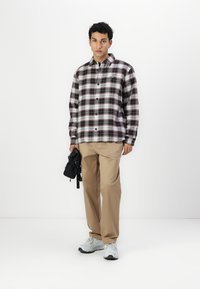 Camisa de franela a cuadros con patrones en rojo oscuro, blanco y negro, abotonada, combinada con pantalones beiges y zapatillas grises, sosteniendo una bolsa negra.