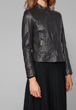 Femme portant une veste en cuir noire cintrée avec des poches à fermeture éclair et boutons-pression, associée à une jupe plissée noire avec des rayures verticales foncées.