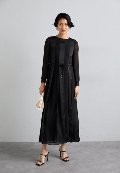 Robe noire à manches longues avec une taille cintrée, fabriquée dans un tissu lisse de type satin. Manches transparentes et ourlet à la cheville. Associée à des talons à brides.