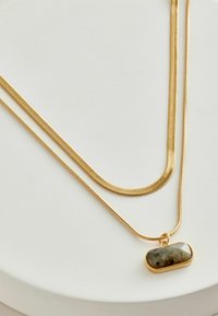 Guldtonade lagerhalsband har eleganta ormkedjor och en hängsmycke med en rundad rektangulär grönaktig sten. Strukturerad yta synlig.