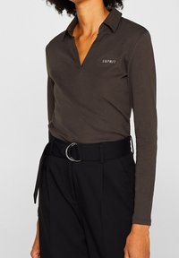 Polo a maniche lunghe marrone scuro con colletto, caratterizzato da un logo con accenti argentati, abbinato a pantaloni neri a vita alta e una cintura.