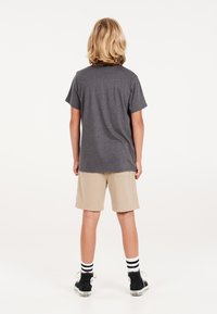 Harmaa puuvillapaita, beige shortsit, mustat korkeakautiset tennarit ja raidalliset sukat, nähtynä takaa valkoista taustaa vasten.