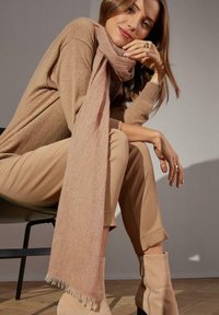 Sciarpa rosa chiaro con bordi sfilacciati che pende su un maglione beige; abbinata a pantaloni beige e stivaletti con dettagli a zip.