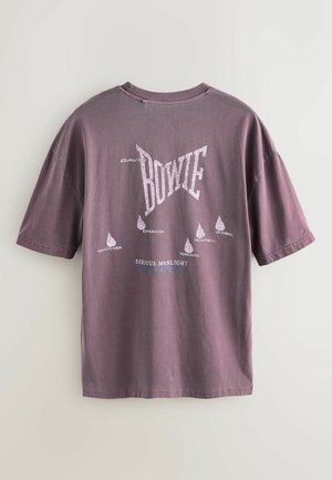 Krátký rukáv, oversize, vybledlé fialové tričko s bílým grafickým textem a místy turné. Měkký bavlněný materiál s volným střihem.