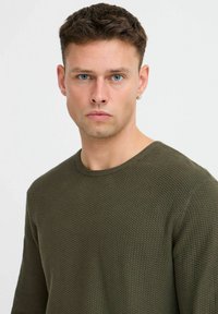 Olivgrüner Strickpullover mit strukturiertem Muster, runder Ausschnitt und langen Ärmeln. Das Modell hat kurze Haare und trägt einen Ohrring.