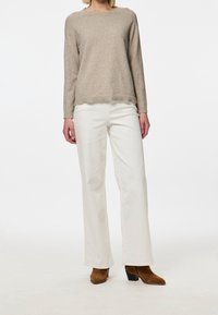 Kvinde iført beige langærmet sweater, hvide vidde bukser og brune hælede ankelstøvler står foran en hvid baggrund.