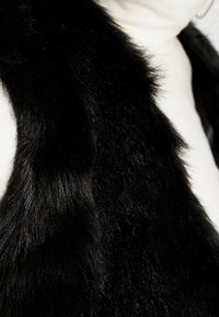 Svart fuskpälsväst med en mjuk, plysjig textur och en slät yta. Har en hög krage och en öppen framdesign.