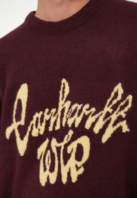 Pull en maille bordeaux avec un texte duveteux crème indiquant « Darkmarks WIP » sur la poitrine, porté par une personne avec un cou et une épaule visibles.