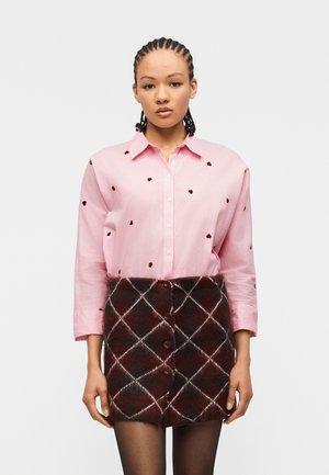 Femme aux cheveux tressés portant une chemise rose à boutons avec des taches rouges et une jupe à carreaux marron boutonnée, debout devant un fond uni.