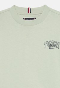 Lys grønn t-skjorte laget av mykt stoff, med rund hals og en liten marineblå "TOMMY HILFIGER"-logo på brystet. Røde og blå detaljer i kragen.
