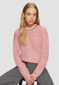 Rosa strukturierte Pullover mit langen Ärmeln und Rundhalsausschnitt, kombiniert mit grauen Hosen. Der Strick hat ein dezentes Muster. 