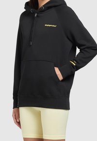 Czarna bluza z kapturem z kieszenią kangurką, z żółtym logo na klatce piersiowej i żółtym akcentem na rękawie. Wykonana z materiału polarowego.