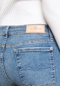 Gros plan sur la poche arrière d'un jean en denim bleu avec un motif de vague brodé et une étiquette de marque en cuir beige, associé à un pull blanc à côtes.