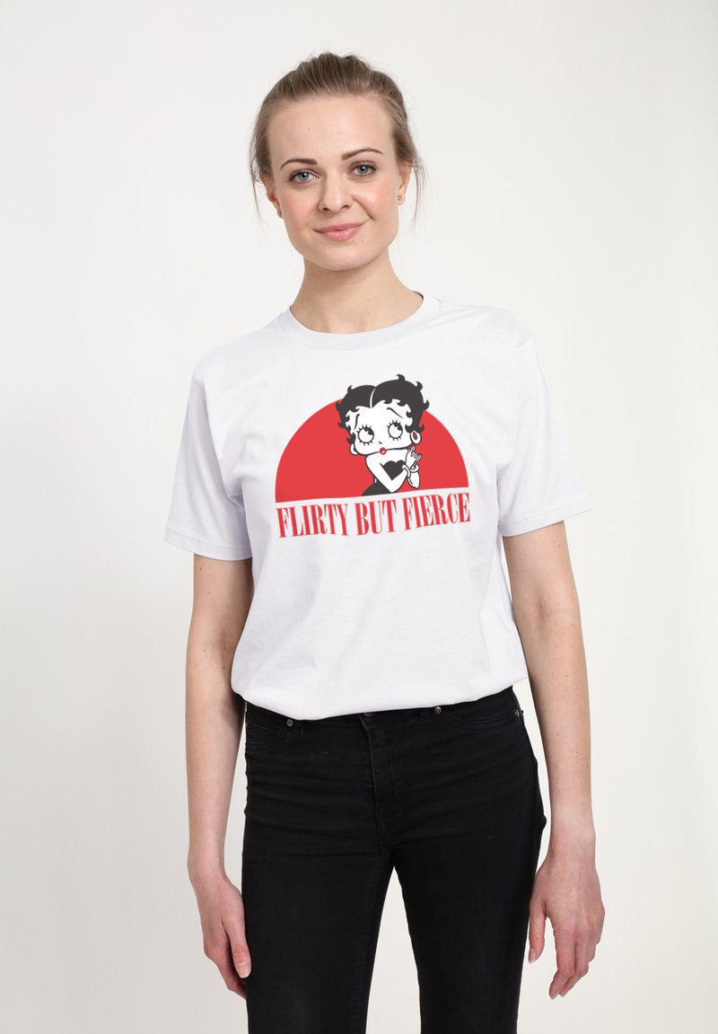 Ung kvinde iført hvid t-shirt med tegneseriefigur og rød tekst "Flirty But Fierce," kombineret med sorte bukser, stående mod en ensfarvet baggrund.