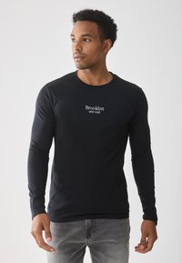 Black long-sleeve shirt with "Brooklyn NEW YORK" printed in white across the chest, made of a smooth, stretchy fabric. Worn with gray jeans.