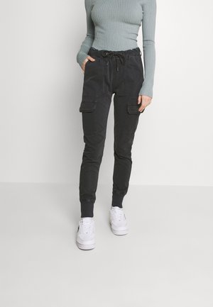 Pantalon cargo noir avec taille à cordon, poignets élastiques, deux poches latérales et coupe droite. Associé à des baskets blanches.