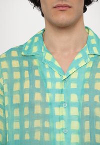 Kortermet skjorte i lett stoff med et turkis og mintgrønt rutenettmønster med gule detaljer og spiss krage.