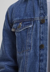 Denim jakna v srednje modri barvi z gumbi na zapiranju, žepom na prsih in kontrastnim rumenim šivom. Mehka, teksturirana tkanina s klasičnim dizajnom.