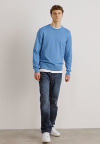 Svetlo modra pulover s okroglim izrezom in rebrastimi manšetami in robom, nošen čez belo majico. Usklajen z temnim denim jeansom in belimi športnimi copati.