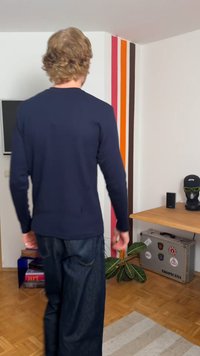 Άνδρας με σγουρά μαλλιά που φοράει ένα ναυτικό πουλόβερ και τζιν, περπατάει μέσα σε ένα δωμάτιο με πολύχρωμες ρίγες στους τοίχους, φυτά και μια βαλίτσα κάτω από ένα ξύλινο γραφείο.