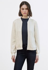 Chaqueta bomber beige claro con cremallera frontal, textura jaspeada, puños y cintura acanalados. Llevada sobre una camiseta oscura ajustada y vaqueros negros.