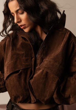 Žena s valovitom tamnom kosom, odjevena u smeđu antilop jaknu s velikim džepovima, gleda dolje sa zatvorenim očima na neutralnoj pozadini.