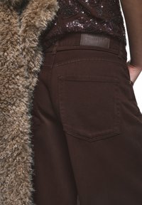 Bruna jeans med en mjuk yta och detalj på bakfickan, kombinerade med en mjuk beige kappa. Lädermärke synligt nära midjan.