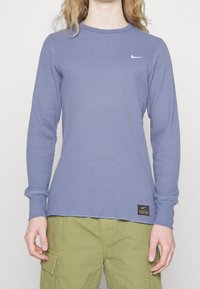 Långärmad, ribbad stickad tröja i dämpad blå färg med rund halsringning och Nike-logotyp. Har en tagg med varumärke vid bottenfåll.