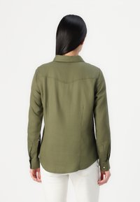 Camicetta verde oliva con colletto, maniche lunghe e orlo curvo. Realizzata in un tessuto morbido con dettagli sulla spalla posteriore.