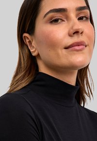 Zwart coltrui met een gladde textuur. Close-up van een gezicht met minimale makeup, met stijlvol bruin haar en gouden oorbellen.