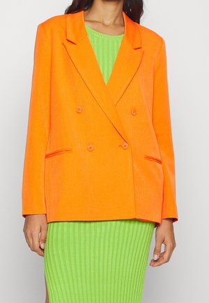 Mujer que lleva un blazer naranja brillante de doble botonadura sobre un vestido de canalé verde claro, de pie contra un fondo claro y liso.