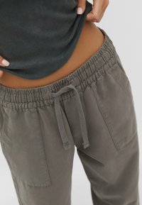 Lähikuva henkilöstä, joka nostaa tumman raidallisen paidan paljastaen vyötärön, jonka päällä on harmaat nyöripöksyt, joissa on etutaskut.